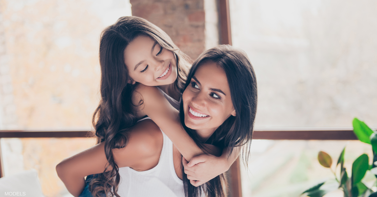 Mother holding her daughter on her back smiling at each other (models)