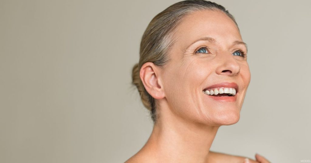 A mature woman with short gray hair smiles warmly at the camera, set against a plain, neutral background (MODEL).