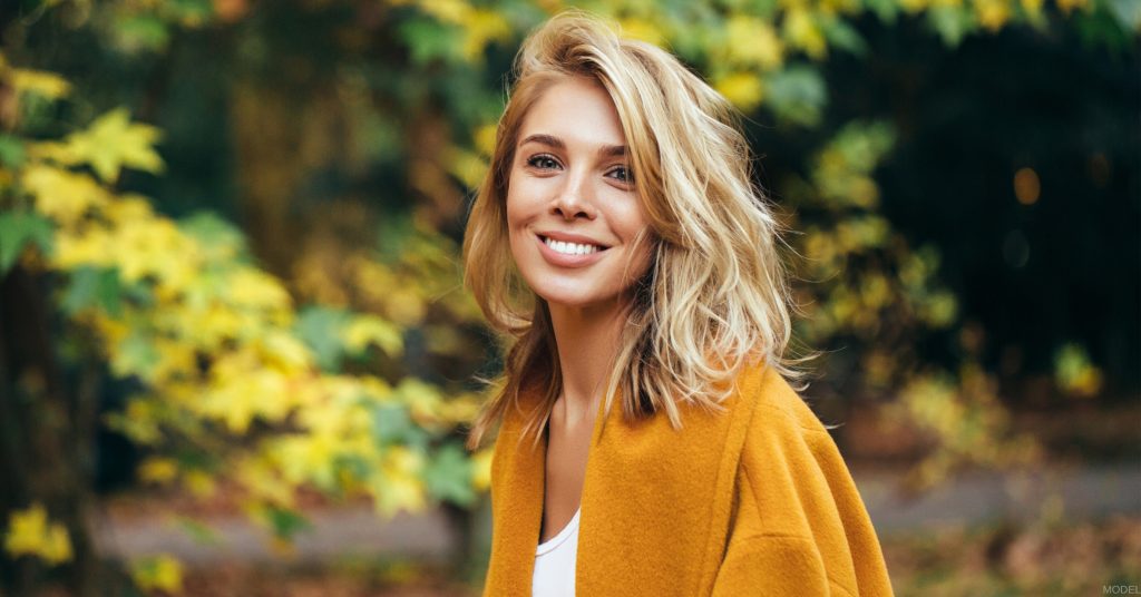 A smiling woman in a yellow cardigan standing in front of autumn trees (model)