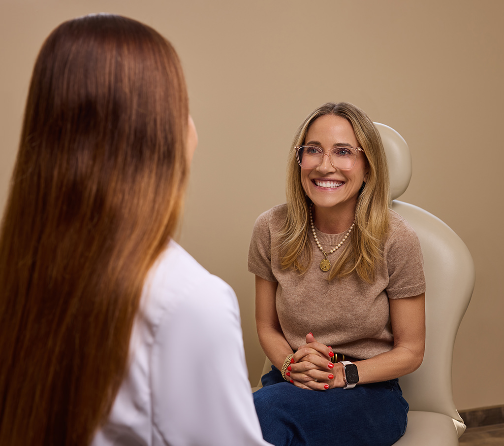 Plastic surgery patient smiling during a consultation with a surgeon in an exam room.